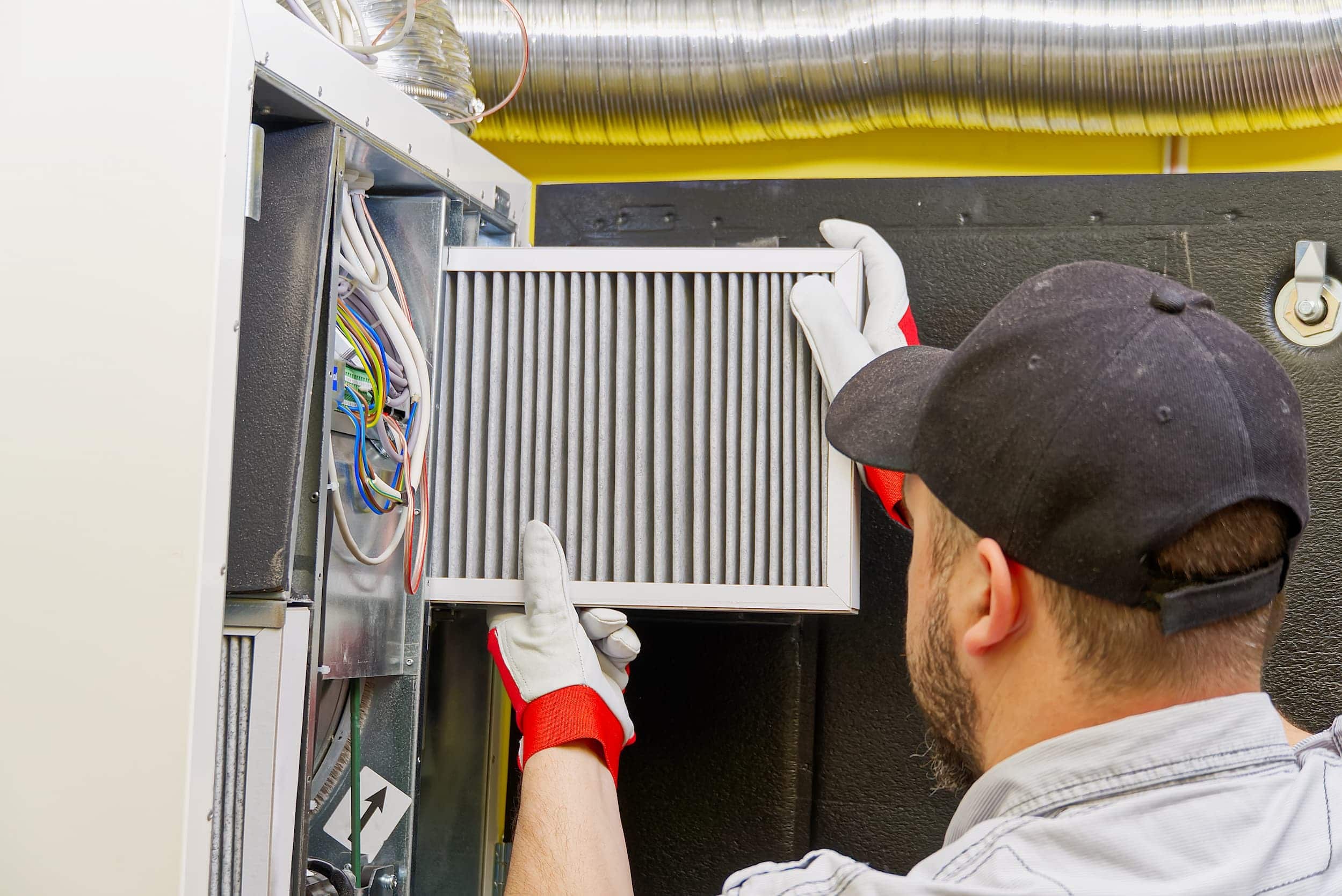 Person changing an air filter in a HVAC system, wearing gloves and a cap, surrounded by wires and ducts. - Southland Mechanical: Industrial Strength HVAC